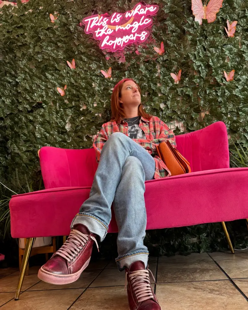 Megan Bresnahan sitting on a pink velvet couch in front of an ivy wall in a burrito shop in San Francisco, underneath a pink neon sign that says 'This is where the magic happens' in cursive.
